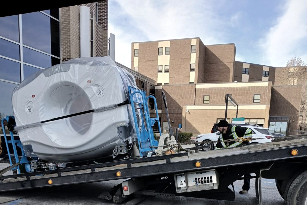 A crew member hooking chains to a shrink-wrapped MRI on a flatbed at a hospital dock