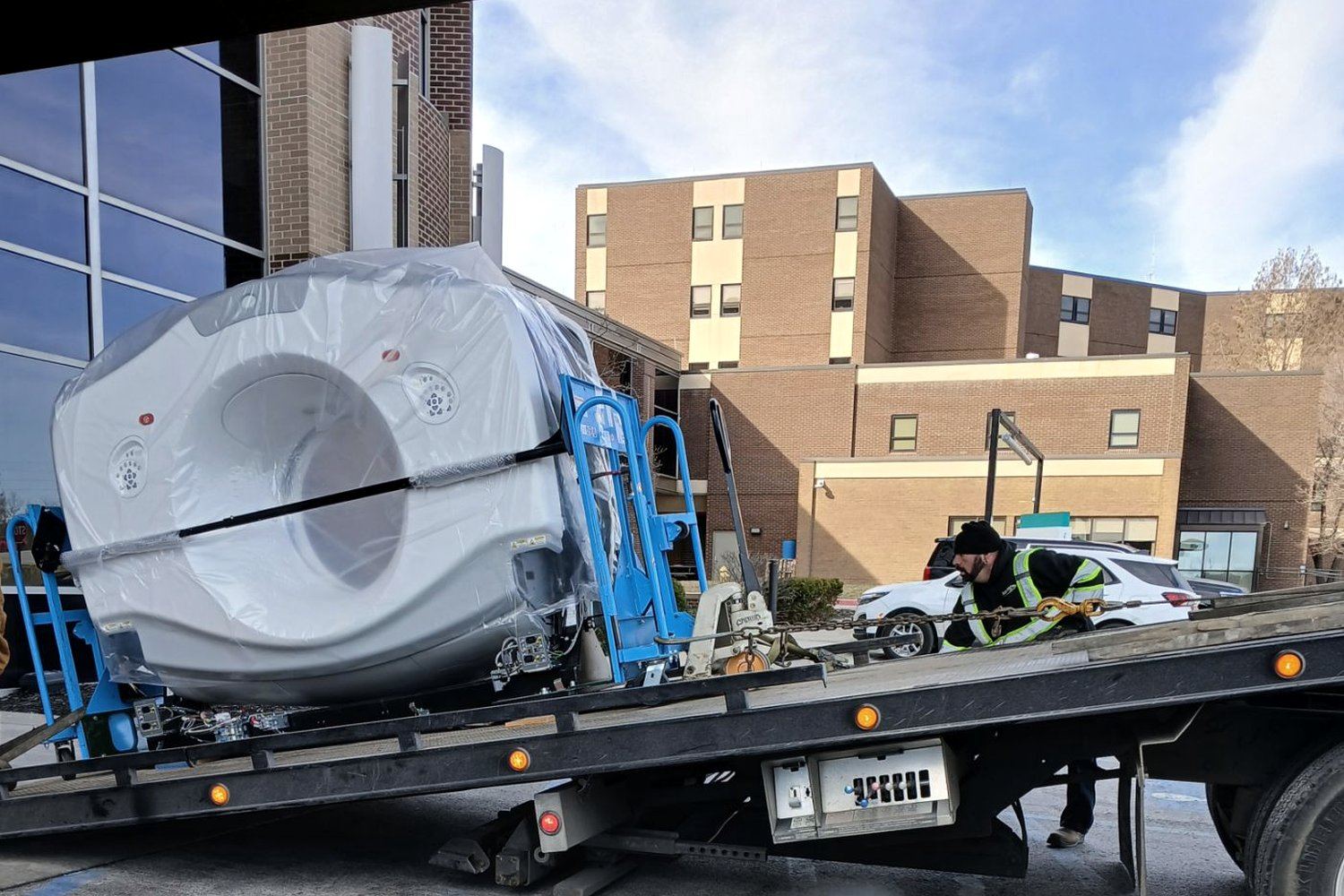 A shrink-wrapped MRI machine on a flatbed outside a hospital while a hi-vis crew member rigs it down with chains